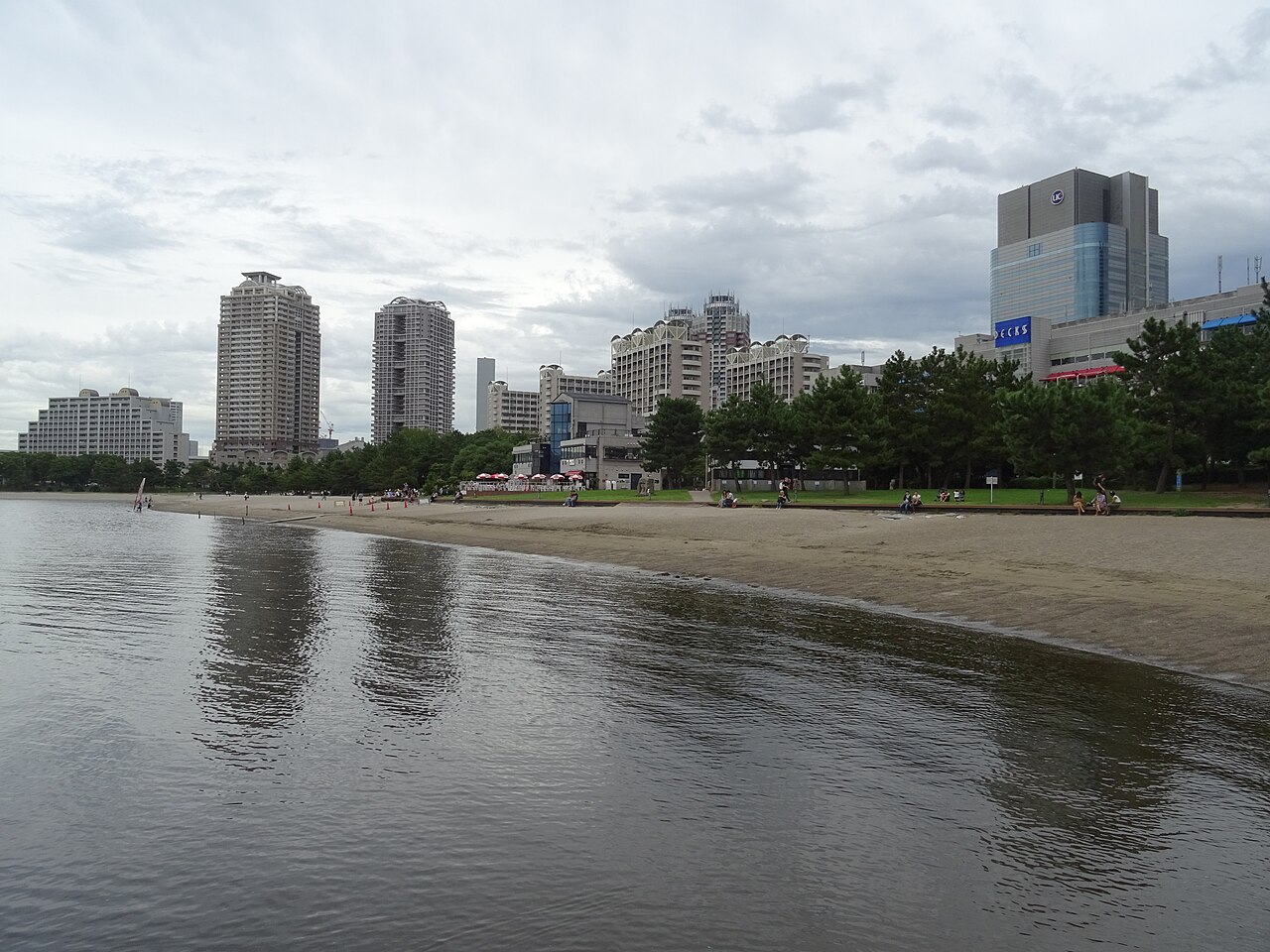 お台場海浜公園の砂浜と水面。奥に並木と台場の建物が並ぶ。