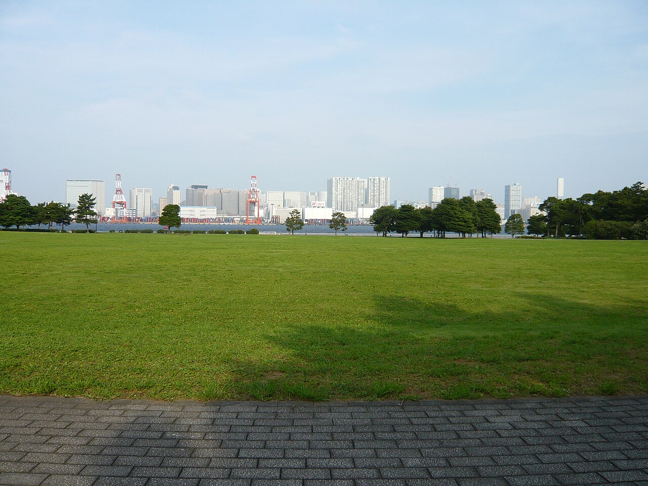潮風公園の広い芝生と木々。海の向こうに港のクレーンと建物が並ぶ。